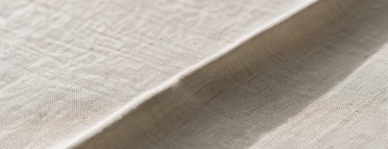Lin crème tendu puis relâché en un seul pli diagonal, texture tissée et ombre portée révélées par une lumière rasante.