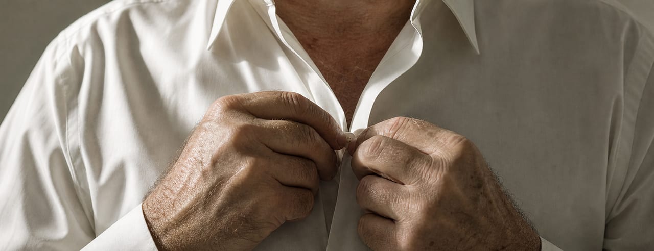 Homme boutonnant une chemise blanche, mains dans le cadre, clavicule visible dans l'ouverture du col, en lumière latérale contrastée.
