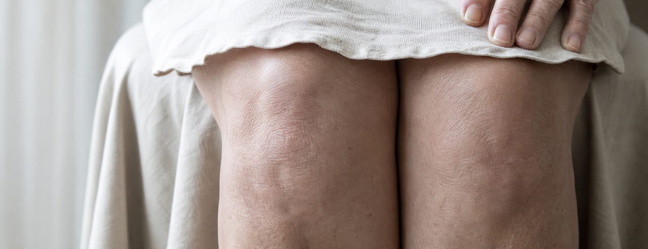 Femme assise, genoux et bas des cuisses jointes, ourlet d'une jupe de lin crème couvrant le haut, en lumière de fenêtre latérale.