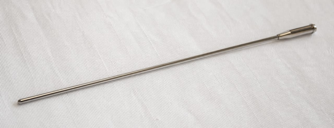 Canule à bout mousse posée seule en diagonale sur un linge chirurgical blanc, fin liseré de lumière sur le métal.