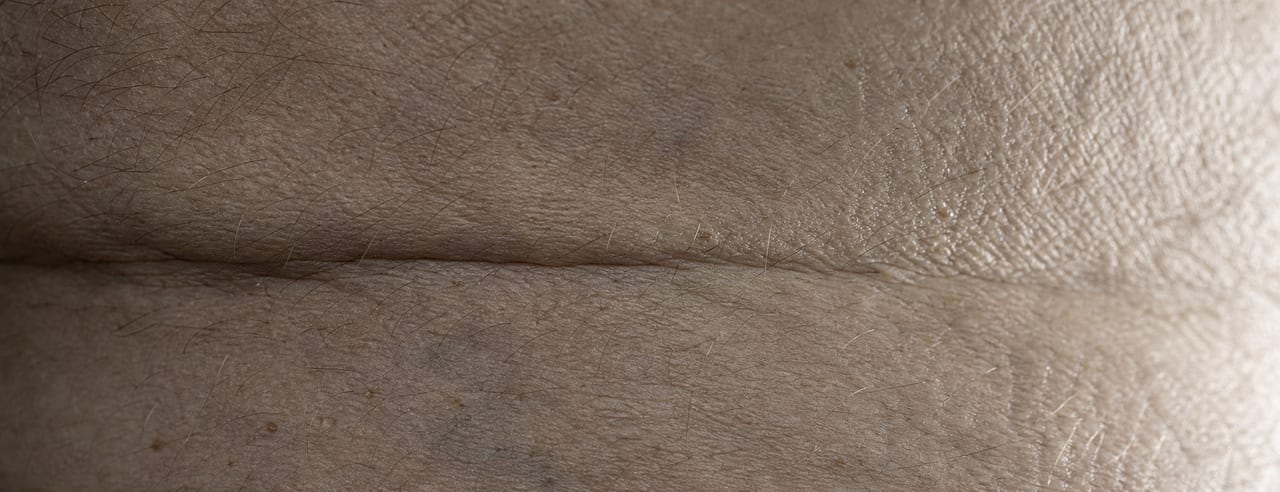 Macro de peau masculine : grain cutané, pilosité fine et un pli naturel traversant le cadre sous une lumière rasante très basse.