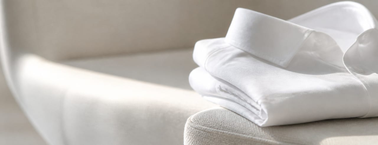 Chemise de coton blanc soigneusement pliée sur le bord d'une chaise claire, traversée par une large lumière de fenêtre.
