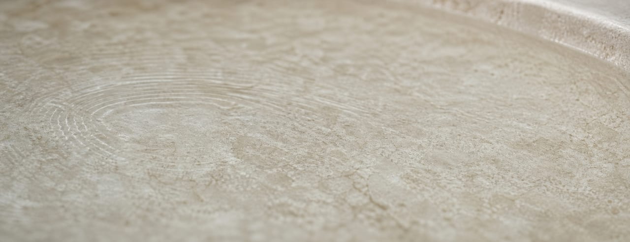 Surface d'eau calme et mate dans une vasque de pierre claire, micro-ondulations concentriques presque éteintes en lumière latérale.
