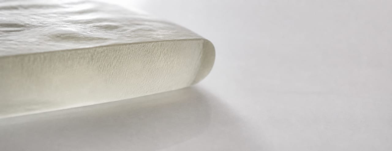 Bord d'un implant en gel de silicone posé sur de la porcelaine blanche, tranche traversée par un contre-jour diffus révélant sa densité.