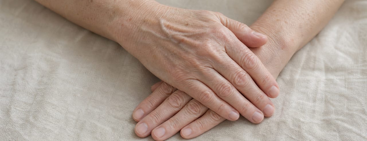 Mains d'une femme âgée posées l'une sur l'autre au repos sur un lin crème, veines, tendons et taches solaires visibles, ongles nus.