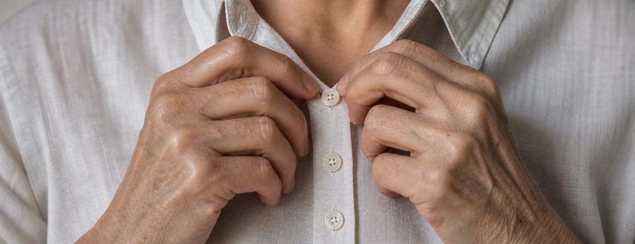Mains d'une femme reboutonnant lentement le haut d'une chemise de lin blanc, boutons de nacre nets à hauteur de clavicule.