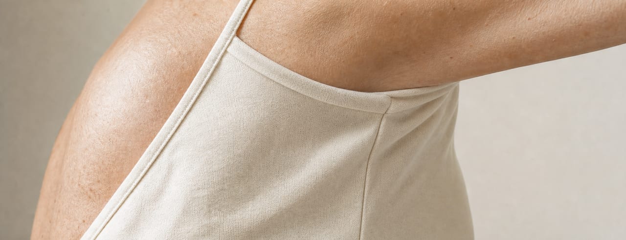 Épaule et flanc d'une femme en débardeur de coton écru, bras à demi levé, textile mat et sec sous une lumière naturelle haute.