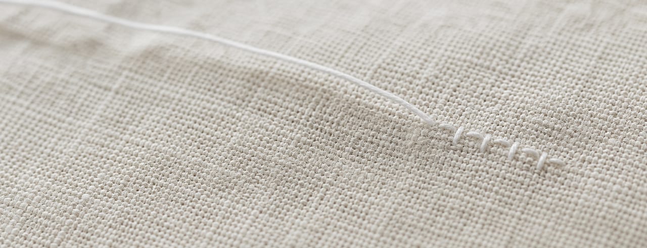 Quelques points de couture main en fil de coton blanc sur une étoffe de lin crème, trame du tissu vibrant en lumière rasante.